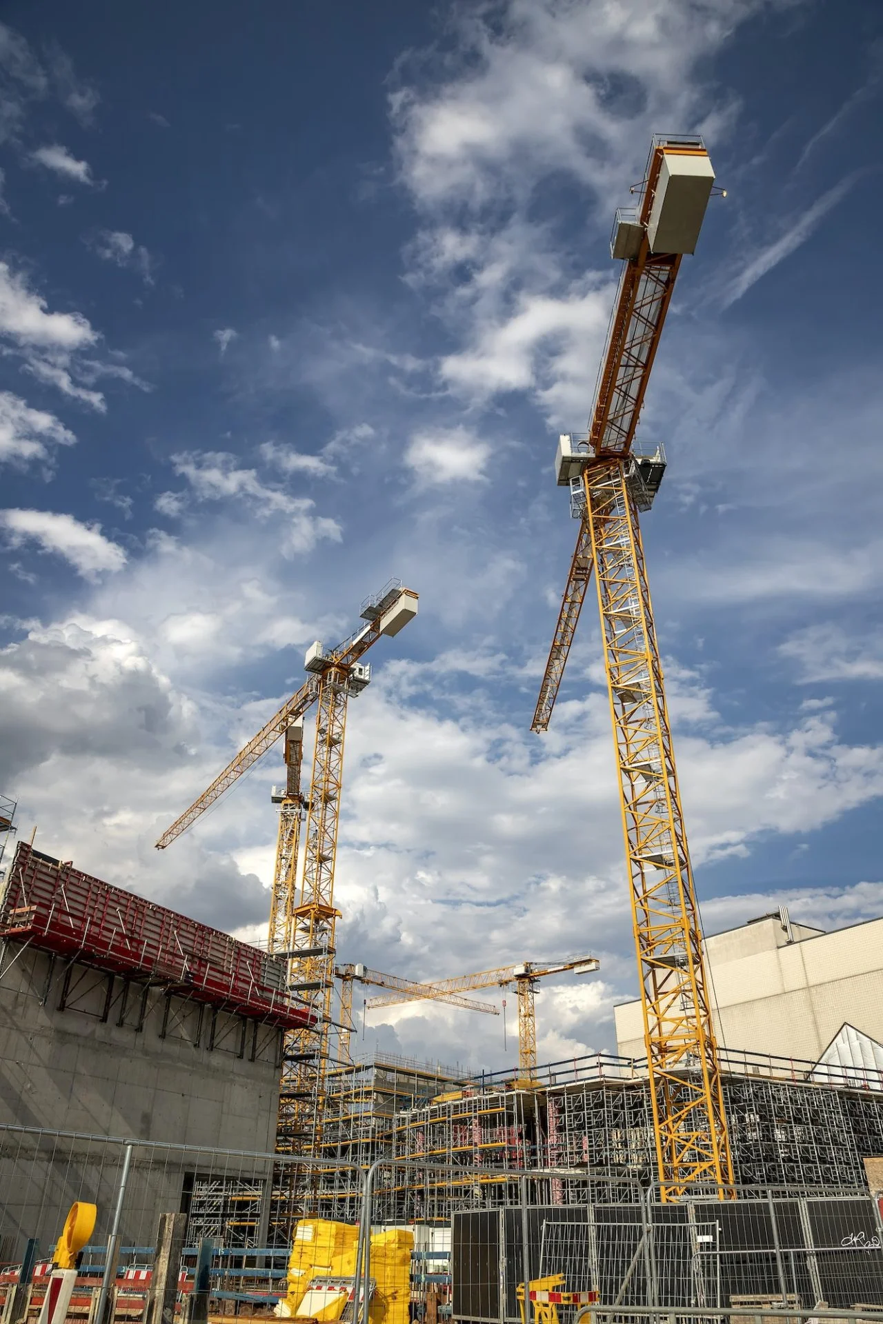 Yellow tower cranes over a concrete building site with red steel formwork, scaffolding, and chain-link fencing