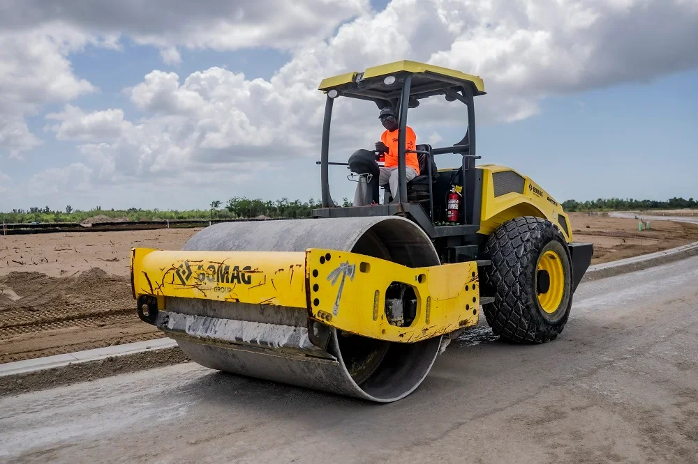 Bomag vibratory soil compactor rolling over graded subgrade with operator in orange high-visibility vest and concrete curb edging